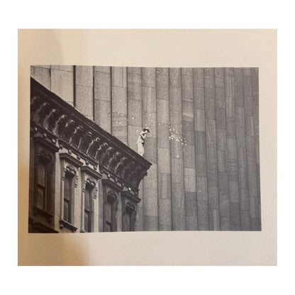 Fotografía en blanco y negro de una figura sobre la cornisa de un edificio junto a una fachada urbana