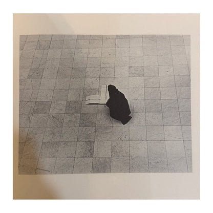 Fotografía en blanco y negro de una figura inclinada junto a un libro abierto sobre suelo de losas