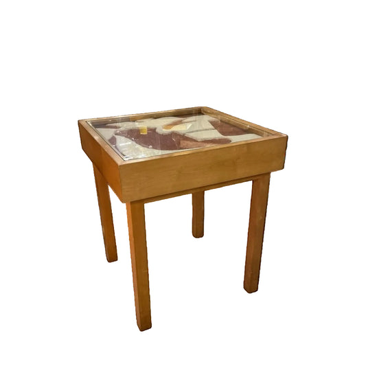 Mesa lateral de madera con estructura de líneas rectas y sobre de cristal protector sobre obra artística