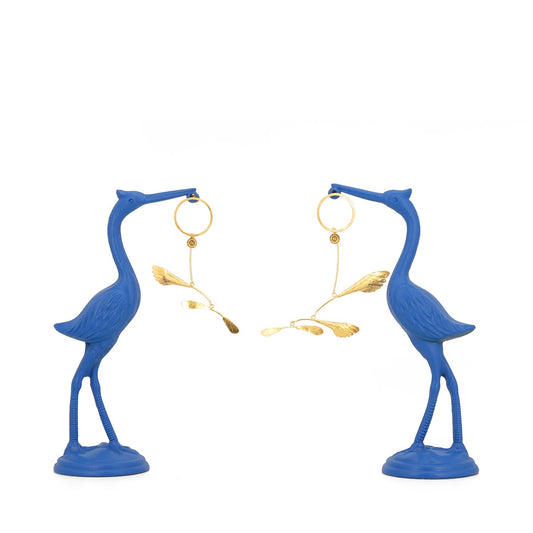 Sculptural gilded silver mobile earrings inspired by Alexander Calder, displayed hanging from two vibrant blue heron statues on a white background
