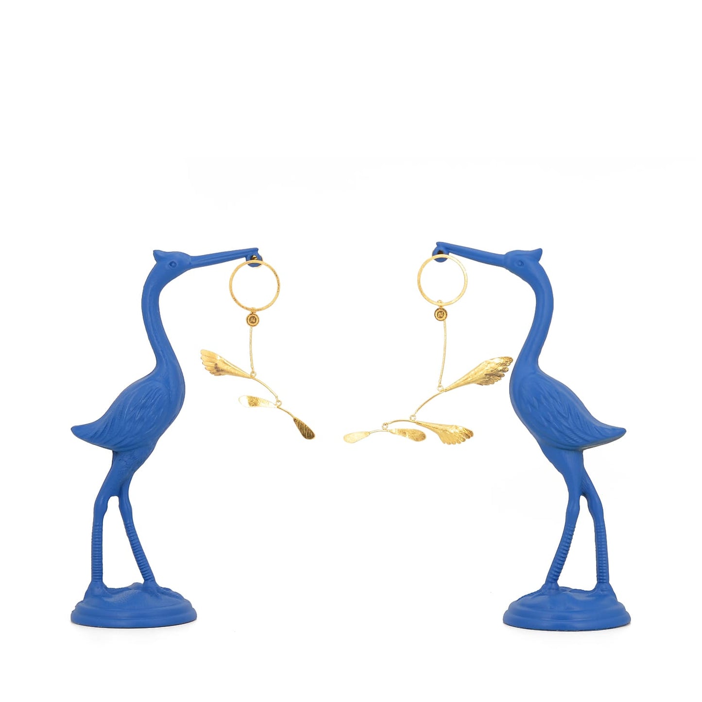 Sculptural gilded silver mobile earrings inspired by Alexander Calder, displayed hanging from two vibrant blue heron statues on a white background