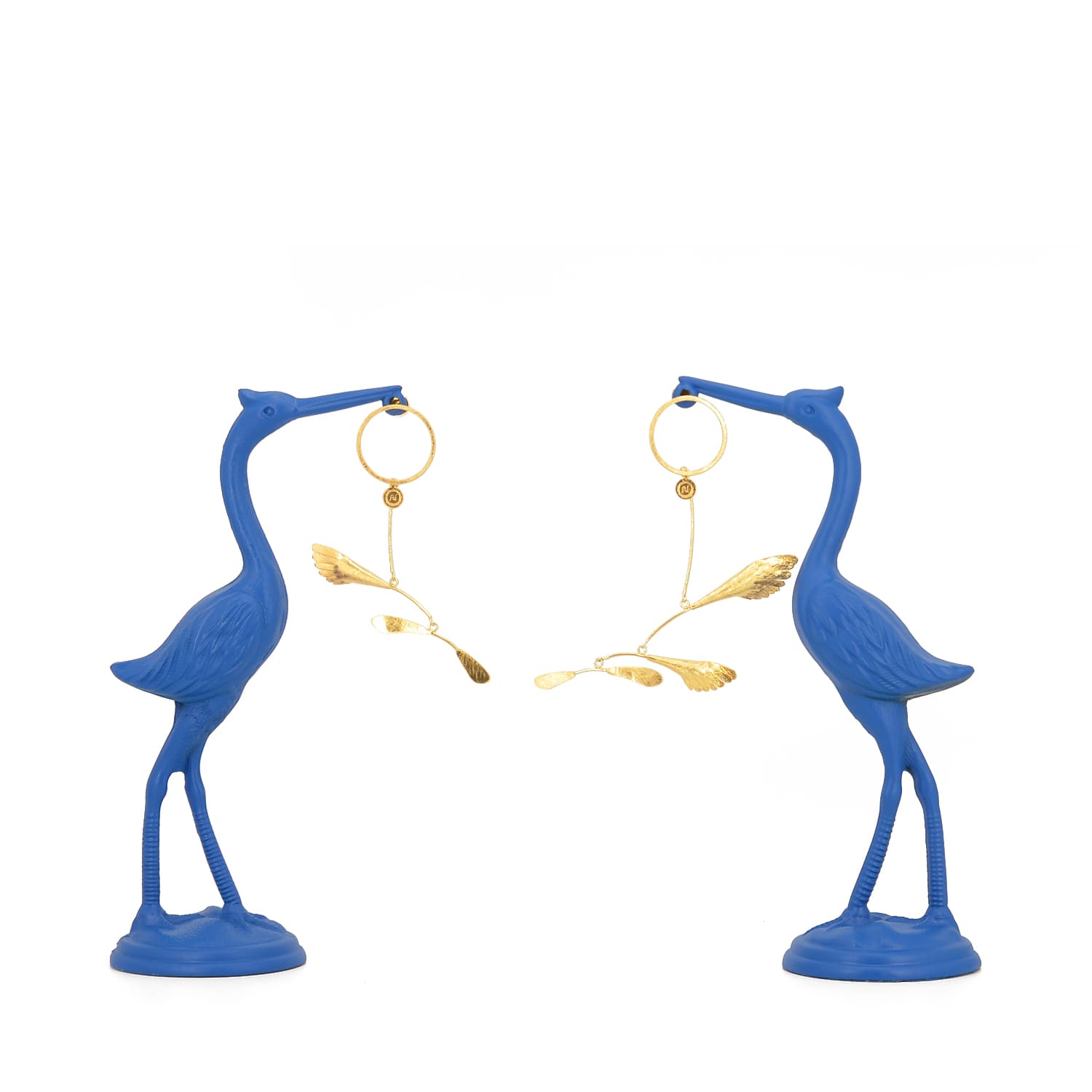 Sculptural gilded silver mobile earrings inspired by Alexander Calder, displayed hanging from two vibrant blue heron statues on a white background
